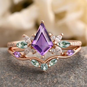 Könnte beinhalten: Ein roségoldenes Ring-Set mit einem großen, rautenförmigen, lila Amethyst-Mittelstein. Der Ring ist mit kleineren weißen und grünen Edelsteinen und einem zarten Band verziert. Das Set enthält ein passendes Band mit kleinen, klaren Steinen.