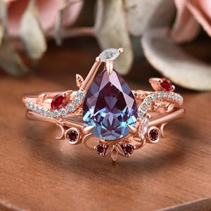 May include: Rose gold ring set featuring a pear-shaped, color-changing alexandrite gemstone. The ring is accented with small red gemstones and white diamonds, with a floral and vine design. The ring is set on a wooden surface.
