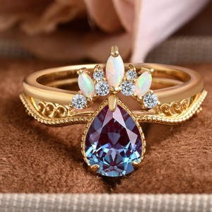 May include: A gold ring featuring a pear-shaped, color-changing alexandrite-style center stone. The band is adorned with a crown-like arrangement of three marquise-cut opals and four small round clear stones, set against a filigree metal band.
