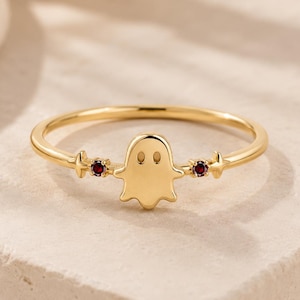 May include: A gold ring featuring a central ghost-shaped charm with two small, round, dark red gemstones set on either side of the ghost figure. The band is thin and polished, showcasing a smooth metallic finish throughout the entire jewelry piece.