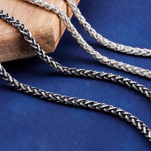 May include: Close-up of three silver wheat chain necklaces. One is dark, oxidised silver, the other two are bright silver. The chains are displayed on a wooden block and a blue surface, showcasing their intricate woven design.