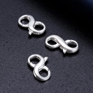 Puede incluir: Tres cierres de infinito plateados. Los cierres están dispuestos sobre una superficie oscura, con un ligero brillo. Los cierres están diseñados para conectar piezas de joyería.