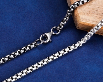 May include: A silver-colored box chain necklace with a lobster clasp. The chain is made of small, square links and is displayed on a blue surface with a wooden accent.