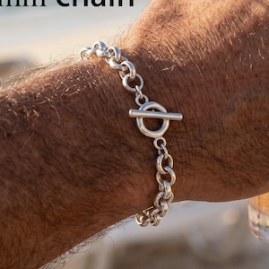 May include: A silver chain bracelet featuring round links and a toggle clasp, with the text 8mm chain visible. The bracelet is constructed from polished silver-toned metal, showcasing a classic link design with a secure bar and ring closure.