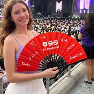 Può includere: Un ventaglio pieghevole rosso con stecche nere che riporta la scritta bianca BTS WORLD TOUR e i nomi Jin, V, J-Hope, Suga, Jimin e Jungkook stampati in bianco. Il ventaglio presenta un logo circolare e stelle su uno sfondo rosso vivo.