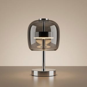 Op de afbeelding: Een moderne tafellamp met een rookglazen kap en een gepolijste chromen voet. De lamp heeft een cilindrische steel en een ronde voet. De kap heeft een afgeronde, klokvormige vorm. De lamp is ontworpen om sfeerverlichting te bieden.