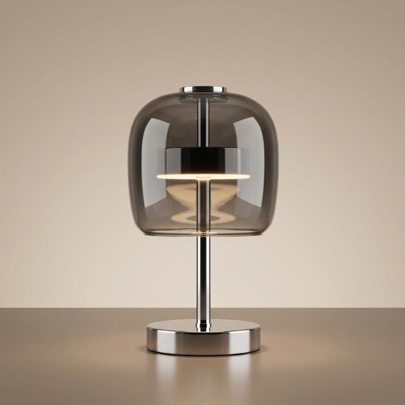 A contemporary minimalist table lamp consisting of a large, rounded-rectangular dome shade made of translucent smoked charcoal grey glass. The dome is supported by a central vertical stem made of polished chrome with a mirror-like silver finish. The lamp exhibits a sleek, architectural design with high-reflectivity on all metal components and a soft, diffused light output that radiates from the center of the dome.