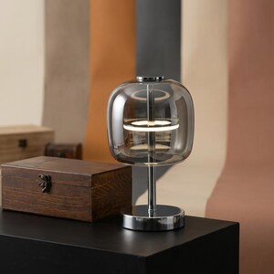 A contemporary minimalist table lamp consisting of a large, rounded-rectangular dome shade made of translucent smoked charcoal grey glass. The dome is supported by a central vertical stem made of polished chrome with a mirror-like silver finish. The lamp exhibits a sleek, architectural design with high-reflectivity on all metal components and a soft, diffused light output that radiates from the center of the dome.