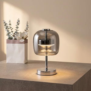 A contemporary minimalist table lamp consisting of a large, rounded-rectangular dome shade made of translucent smoked charcoal grey glass. The dome is supported by a central vertical stem made of polished chrome with a mirror-like silver finish. The lamp exhibits a sleek, architectural design with high-reflectivity on all metal components and a soft, diffused light output that radiates from the center of the dome.