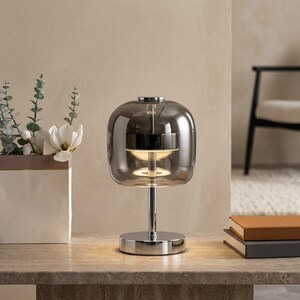A contemporary minimalist table lamp consisting of a large, rounded-rectangular dome shade made of translucent smoked charcoal grey glass. The dome is supported by a central vertical stem made of polished chrome with a mirror-like silver finish. The lamp exhibits a sleek, architectural design with high-reflectivity on all metal components and a soft, diffused light output that radiates from the center of the dome.