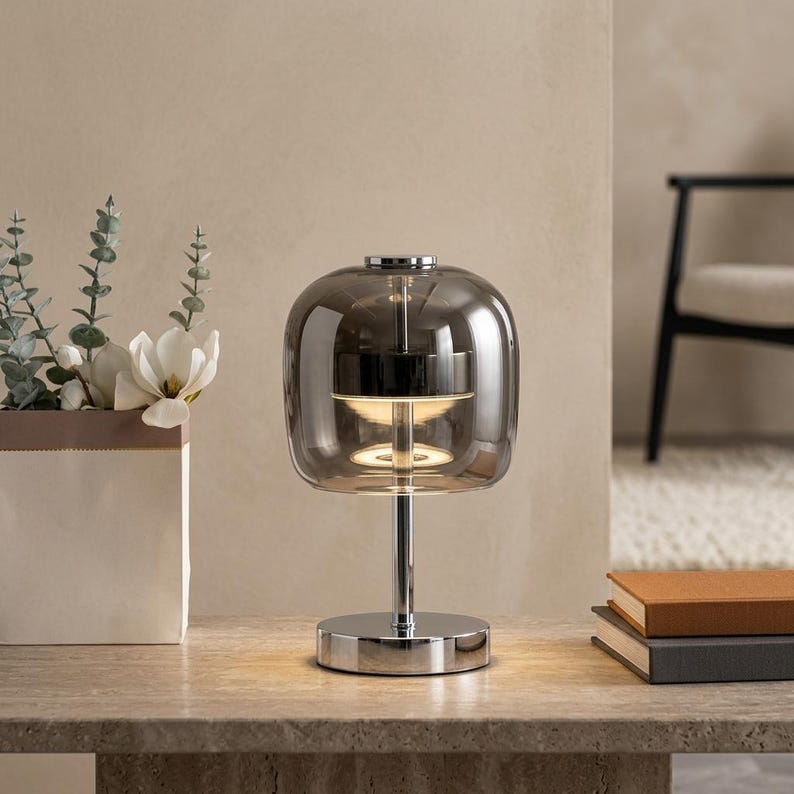 A contemporary minimalist table lamp consisting of a large, rounded-rectangular dome shade made of translucent smoked charcoal grey glass. The dome is supported by a central vertical stem made of polished chrome with a mirror-like silver finish. The lamp exhibits a sleek, architectural design with high-reflectivity on all metal components and a soft, diffused light output that radiates from the center of the dome.