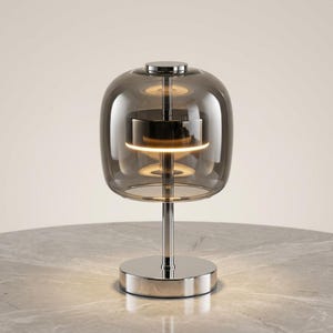 A contemporary minimalist table lamp consisting of a large, rounded-rectangular dome shade made of translucent smoked charcoal grey glass. The dome is supported by a central vertical stem made of polished chrome with a mirror-like silver finish. The lamp exhibits a sleek, architectural design with high-reflectivity on all metal components and a soft, diffused light output that radiates from the center of the dome.