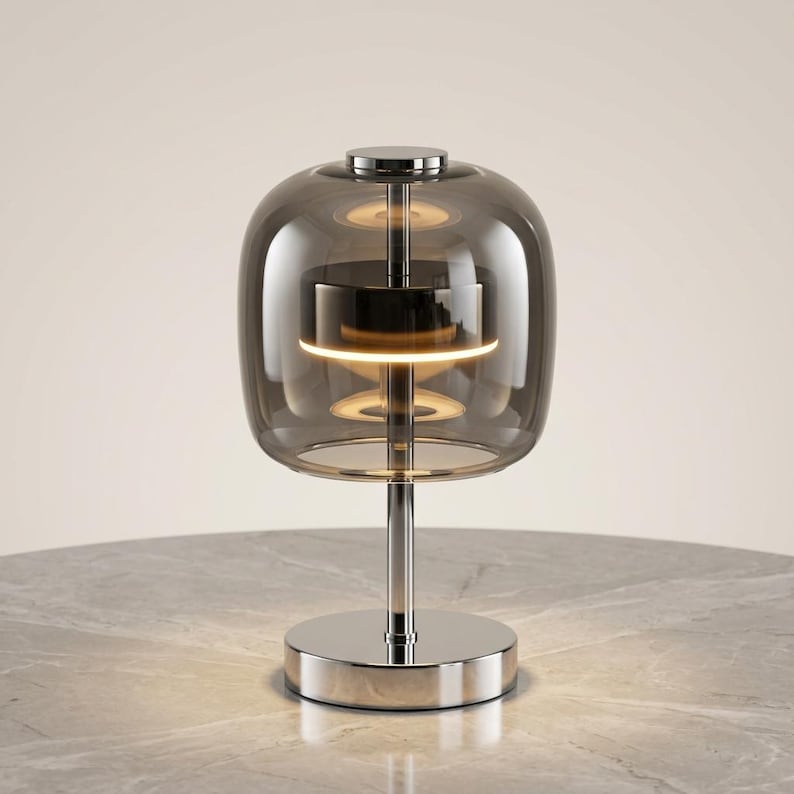 A contemporary minimalist table lamp consisting of a large, rounded-rectangular dome shade made of translucent smoked charcoal grey glass. The dome is supported by a central vertical stem made of polished chrome with a mirror-like silver finish. The lamp exhibits a sleek, architectural design with high-reflectivity on all metal components and a soft, diffused light output that radiates from the center of the dome.