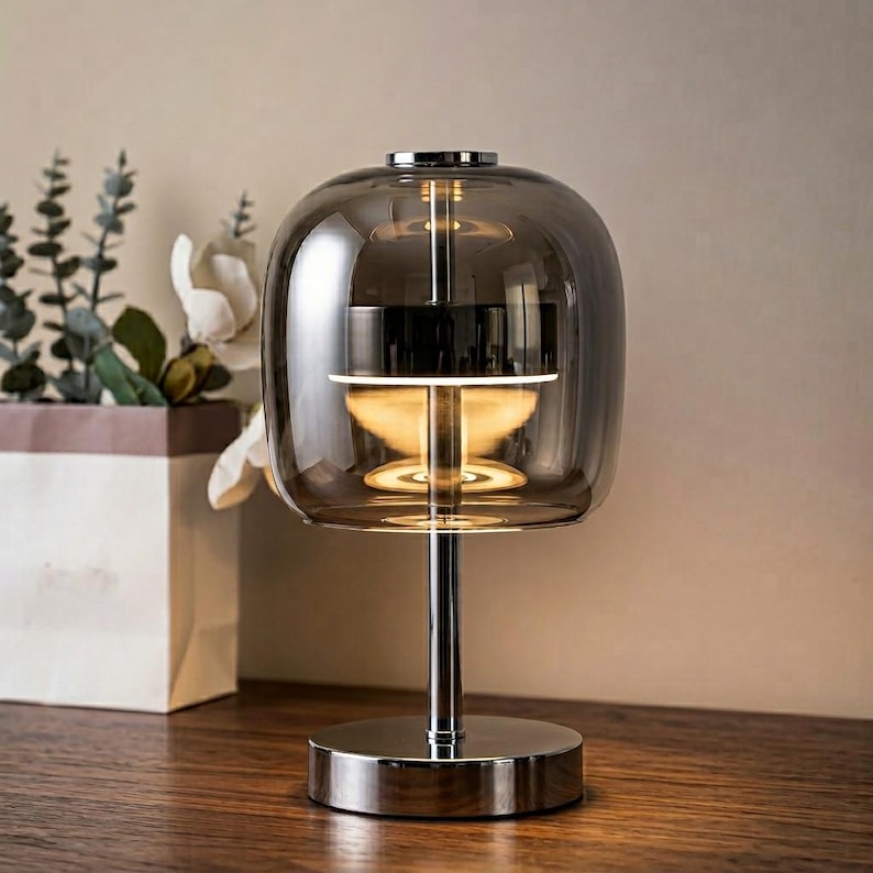 A contemporary minimalist table lamp consisting of a large, rounded-rectangular dome shade made of translucent smoked charcoal grey glass. The dome is supported by a central vertical stem made of polished chrome with a mirror-like silver finish. The lamp exhibits a sleek, architectural design with high-reflectivity on all metal components and a soft, diffused light output that radiates from the center of the dome.