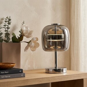 A contemporary minimalist table lamp consisting of a large, rounded-rectangular dome shade made of translucent smoked charcoal grey glass. The dome is supported by a central vertical stem made of polished chrome with a mirror-like silver finish. The lamp exhibits a sleek, architectural design with high-reflectivity on all metal components and a soft, diffused light output that radiates from the center of the dome.