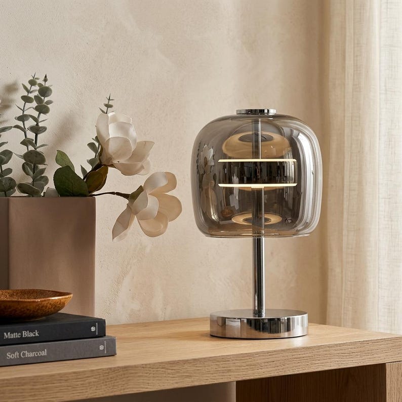 A contemporary minimalist table lamp consisting of a large, rounded-rectangular dome shade made of translucent smoked charcoal grey glass. The dome is supported by a central vertical stem made of polished chrome with a mirror-like silver finish. The lamp exhibits a sleek, architectural design with high-reflectivity on all metal components and a soft, diffused light output that radiates from the center of the dome.