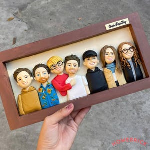 Può includere: Un ritratto di famiglia incorniciato con sette figure scolpite, ognuna con tratti del viso e abbigliamento distinti. Le figure sono disposte in fila all'interno di una cornice di legno marrone. Il testo "Our Family" è visibile sulla cornice.