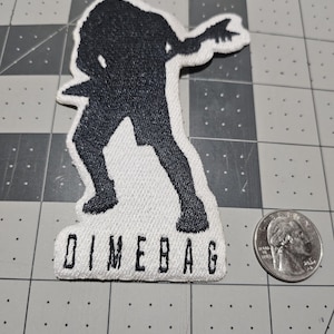 May include: A white embroidered patch featuring a black silhouette of a person playing an electric guitar. The text DIMEBAG is stitched in black capital letters at the bottom. A US quarter is placed next to the patch for scale reference.