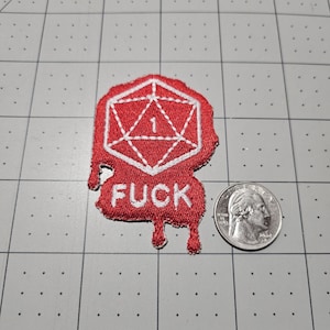 May include: A red embroidered patch featuring a white outline of a twenty-sided die with the number 1 inside and the word FUCK below it, with a dripping effect at the bottom. A US quarter coin is placed next to the patch for scale reference.