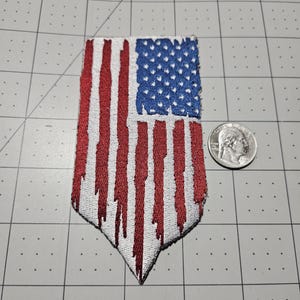 May include: A vertical embroidered patch featuring a stylized American flag design with red and white stripes that appear to drip downward, alongside a blue field with white stars. A silver United States quarter is placed next to the patch for scale.