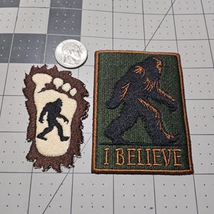 May include: Two embroidered patches featuring a Bigfoot theme. One patch is a cream-colored footprint with a black Bigfoot silhouette, and the other is a rectangular olive green patch with a black Bigfoot figure and the text I BELIEVE in orange.