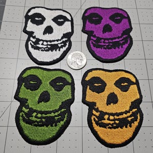 May include: Four embroidered skull patches in white, purple, green, and yellow, each featuring black details and a black border. A silver United States dime is placed in the center for scale, showing the relative size of the patches.