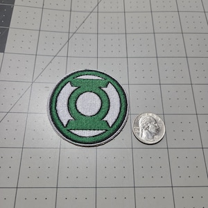 May include: A circular embroidered patch featuring the green and white Green Lantern logo. The patch is positioned next to a standard United States quarter coin for scale, showing the relative size of the circular emblem on a grid mat.