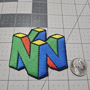 May include: An embroidered patch featuring the N64 logo in blue, green, red, and yellow, with a black border. The patch is positioned next to a standard U.S. quarter coin for scale, showing the patch is approximately 2.5 inches in width.