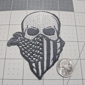 May include: An embroidered patch featuring a skull wearing a bandana patterned with the American flag. The patch is shown next to a United States quarter, which has a diameter of approximately 0.955 inches, for scale on a cutting mat.
