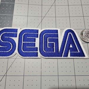 May include: A blue and white embroidered patch featuring the word SEGA in a bold, stylized font. The patch is positioned next to a silver United States quarter coin for scale, which measures approximately 0.955 inches in diameter on the grid surface.