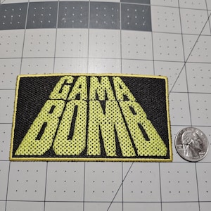 May include: A rectangular embroidered patch featuring the text GAMA BOMB in bold, yellow, block-style lettering against a black background with a yellow border. A silver US quarter coin is placed to the right for scale on a grid mat.