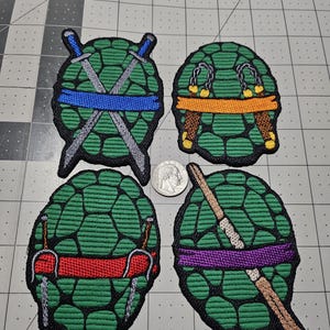 May include: Four embroidered patches shaped like green turtle shells, each featuring a different colored mask and weapon: blue with swords, orange with nunchucks, red with sai, and purple with a bo staff. A US quarter coin is placed in the center.