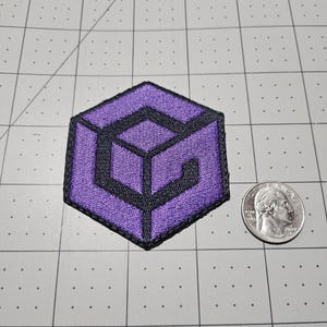 May include: A hexagonal embroidered patch featuring a purple and black geometric design resembling a stylized letter G. The patch is positioned next to a standard United States quarter coin for scale, showing the relative size of the embroidered item.