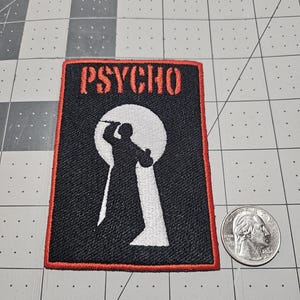 May include: A rectangular embroidered patch with a black background and a red border. It features the word PSYCHO in red letters at the top and a white silhouette of a figure holding a knife inside a keyhole shape. A US quarter is shown for scale.