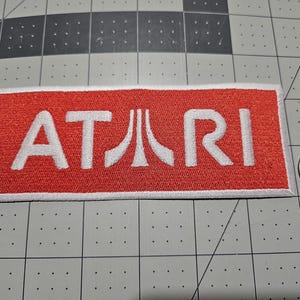 May include: A rectangular embroidered patch featuring the white Atari logo on a red background with a white border. The patch is positioned next to a silver United States quarter coin for scale, showing the relative size of the embroidered item.