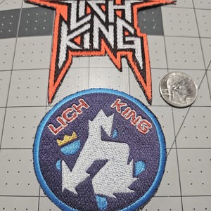 May include: Two embroidered patches featuring the text Lich King. The top patch is a jagged, orange-bordered logo. The bottom patch is circular with a purple background, a white stylized symbol, a small yellow crown, and a US quarter for scale.