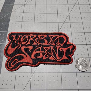 May include: A black embroidered patch with red stitching featuring the text "MORBID SAINT" in a stylized, gothic-inspired font. The patch is positioned next to a United States quarter coin for scale, which measures approximately 0.955 inches in diameter.