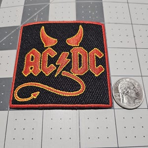 May include: A rectangular embroidered patch featuring the black AC/DC logo with red and yellow outlines. The design includes red devil horns above the text and a red devil tail curving below. A US quarter is placed next to the patch for scale.