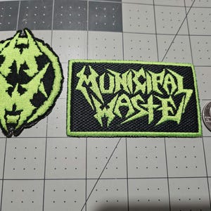 May include: Two embroidered patches with black backgrounds and lime green stitching. One patch is circular with a stylized logo, and the other is rectangular with the text MUNICIPAL WASTE in a jagged font. A US quarter coin is shown for scale.
