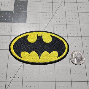 May include: An oval-shaped embroidered patch featuring a black bat silhouette centered on a yellow background with a black border. The patch is positioned next to a standard U.S. quarter coin for scale, providing a clear visual reference for its size.