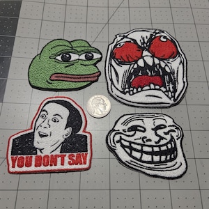 May include: Four embroidered patches featuring internet memes: a green frog, a red-eyed screaming face, a man with the text "YOU DON'T SAY," and a black-and-white troll face. A US quarter coin is placed in the center for scale reference.