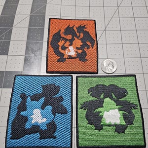 May include: Three rectangular embroidered patches featuring stylized creature silhouettes. One patch is orange with a dark charcoal design, one is blue with a dark charcoal design, and one is green with a dark charcoal design. A US quarter is shown for scale.