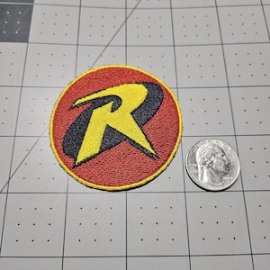 May include: A circular embroidered patch featuring a stylized yellow letter R on a black background, surrounded by a red field and a yellow border. The patch is positioned next to a standard United States quarter coin for size reference.