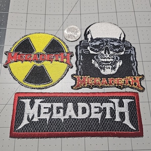 May include: Three embroidered fabric patches featuring the Megadeth logo and imagery. One circular yellow and black radiation symbol patch, one skull character patch, and one rectangular black patch with white text. A US quarter is shown for scale.