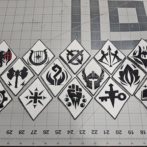 May include: A collection of fourteen diamond-shaped embroidered patches featuring various black and white symbols, including a dragon with red DM text, a harp, swords, a helmet, a key, and a book, arranged on a cutting mat with a US quarter coin.