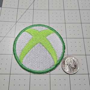 May include: A circular embroidered patch featuring a lime green X shape centered on a light gray background with a lime green border. The patch is positioned next to a United States quarter coin for scale, showing the patch is approximately 2 inches wide.
