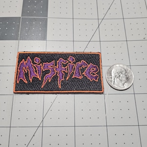 May include: A rectangular embroidered patch featuring the word Misfire in purple thread with a jagged, stylized font on a black background, bordered by an orange stitched edge, shown next to a standard U.S. quarter for scale reference.