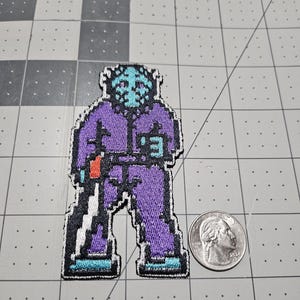 May include: An embroidered patch featuring a pixelated character wearing a light blue hockey mask and a purple jumpsuit. The character holds a white blade with an orange handle. A US quarter is placed next to the patch for scale reference.