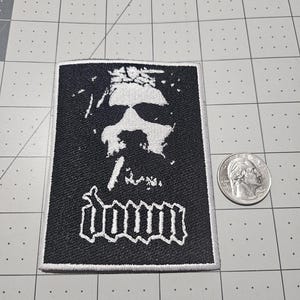 May include: A rectangular black and white embroidered patch featuring a stylized graphic of a face and the word "down" in a gothic font. A US quarter coin is placed next to the patch for scale on a grid-patterned cutting mat surface.