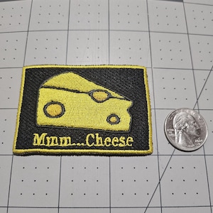 May include: A rectangular embroidered patch with a black background and a yellow border. The patch features a yellow Swiss cheese wedge graphic and the text "Mmm...Cheese" in yellow embroidery, shown next to a US quarter for scale.